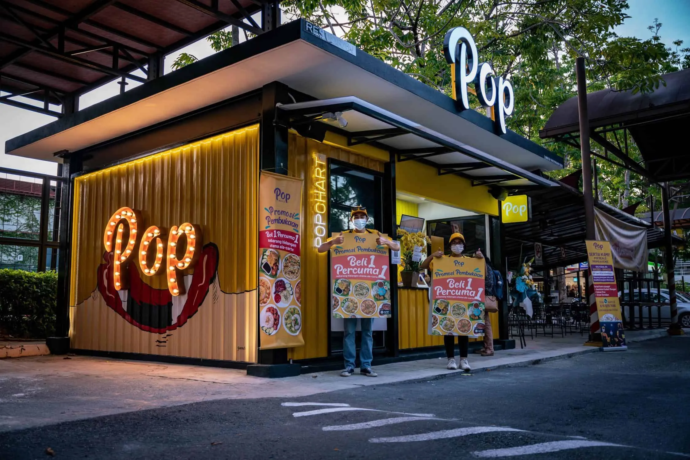 A picture of an outdoor outlet of Pop Meals with two staff standing outside holding signs promoting a new buy 1 free 1 deal